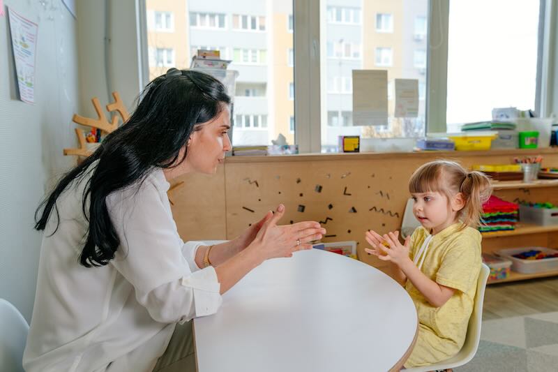 Small kindergarten-age girl sitting at a table with a speech therapist and working with her while several sensory toys are strewn around