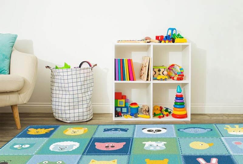 A Playroom Rug with various animals in colorful squares in a kids room along with a white wooden bookcase with books and some colorful sensory toys on the shelf. There is also a kids chair and a toy sack on the floor. 