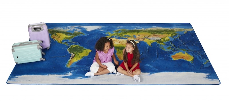 2 kids seating on a colorful classroom rug that has printed a realistic map of Earth on it with 2 suitcases lying around in the upper left corner